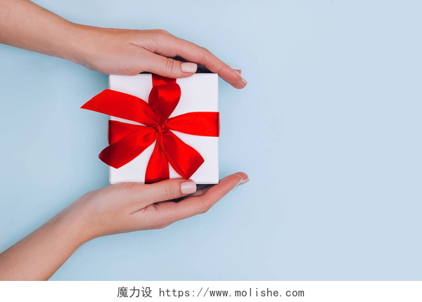蓝色背景上手拿礼物盒子女性的手与指甲保持礼品盒与红丝带淡蓝色背景。 节日的概念.